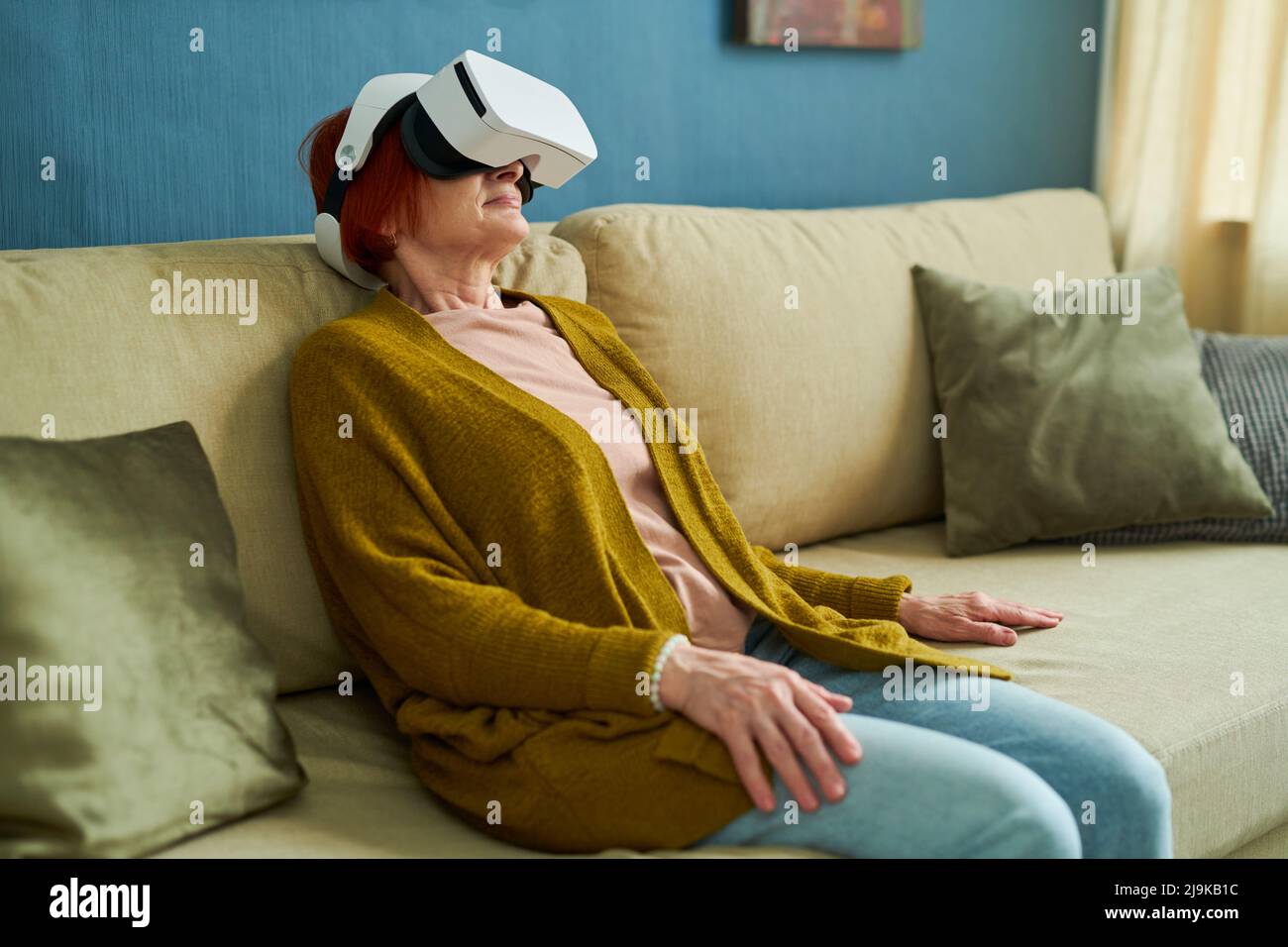 Ältere Frau in VR-Brille mit Virtual Reality zum Entspannen, während sie auf dem Sofa im Wohnzimmer sitzt Stockfoto