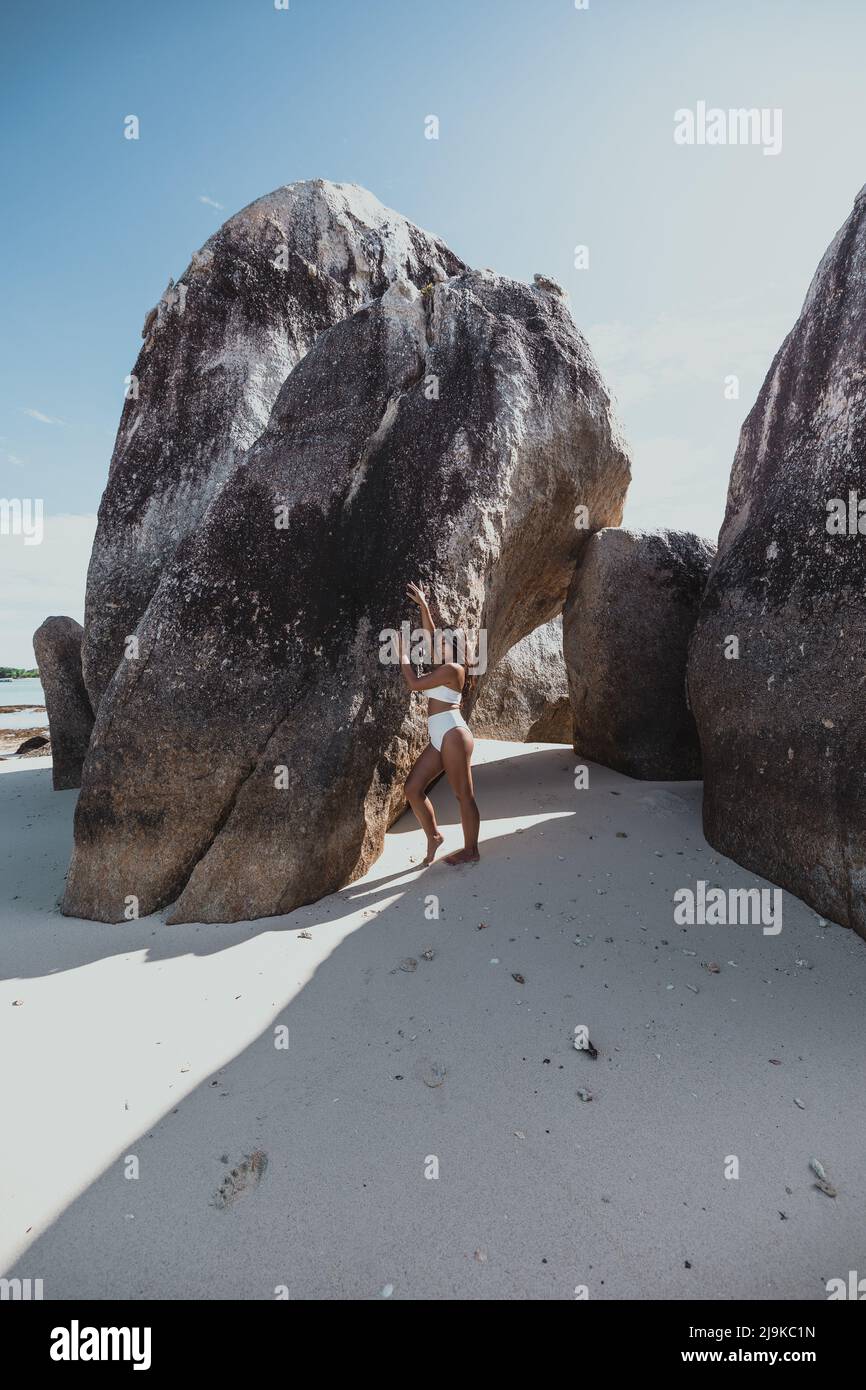 Junge, braune asiatische Frau, die an einem weißen tropischen Sandstrand in Belitung Indonesia neben riesigen Felsen steht Stockfoto