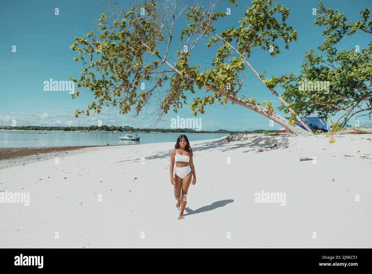 Die junge, bräunlich braune asiatische Frau verlassene an einem weißen Sandstrand in einem weißen Bikini-Badeanzug mit einem blauen Zelt auf der Insel und einem Baum, der über sie fiel Stockfoto