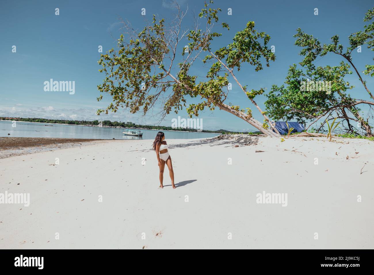 Die junge, bräunlich braune asiatische Frau verlassene an einem weißen Sandstrand in einem weißen Bikini-Badeanzug mit einem blauen Zelt auf der Insel und einem Baum, der über sie fiel Stockfoto