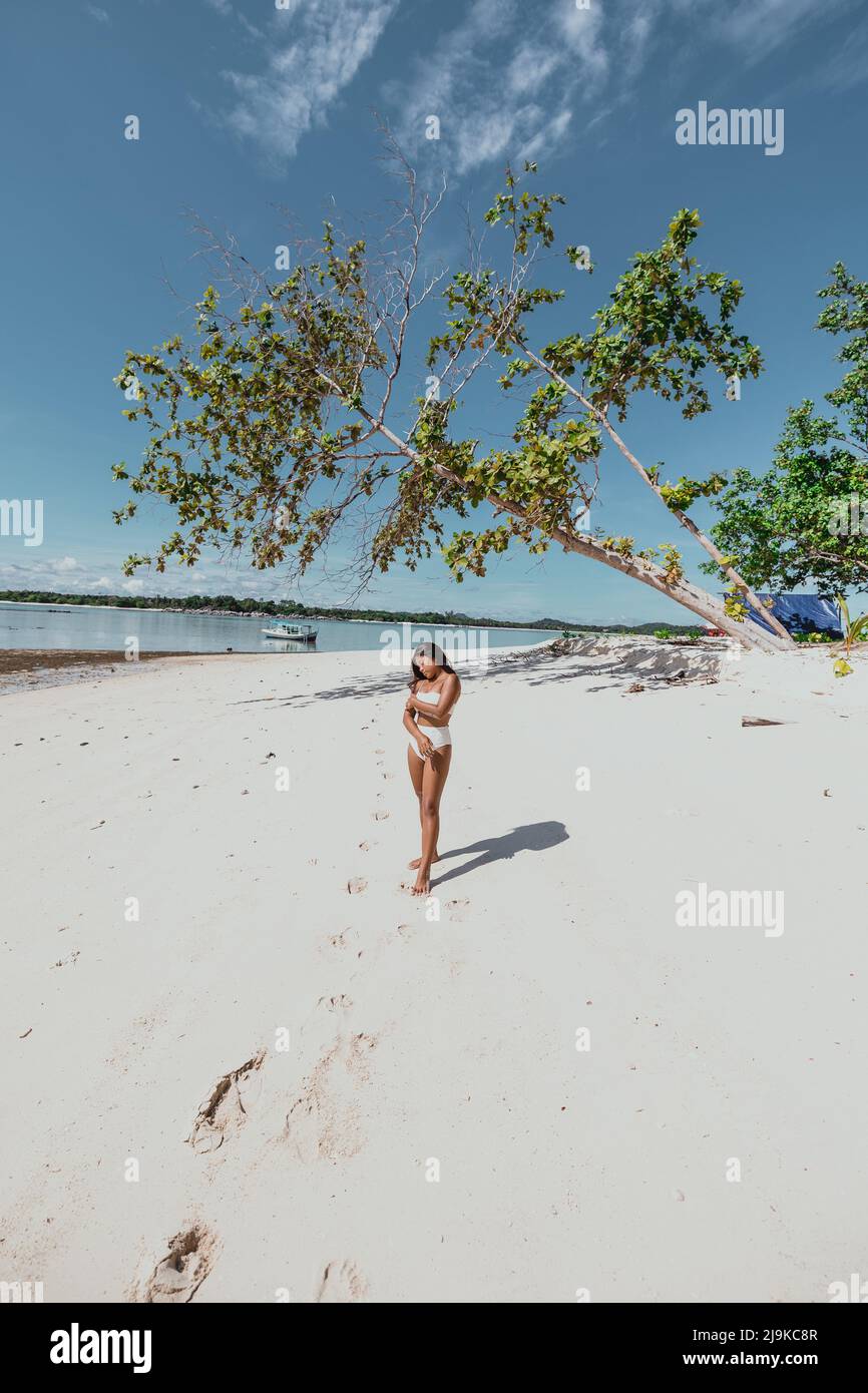 Die junge, bräunlich braune asiatische Frau verlassene an einem weißen Sandstrand in einem weißen Bikini-Badeanzug mit einem blauen Zelt auf der Insel und einem Baum, der über sie fiel Stockfoto