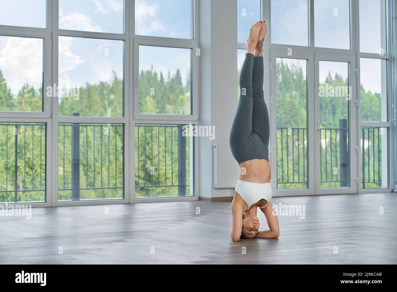 Gesunde, fit Frau beim Training am Handstand in der geräumigen Halle mit Panoramafenstern. Junge Dame in Sportkleidung praktiziert Yoga im Haus. Gesunde Lebensweise. Stockfoto
