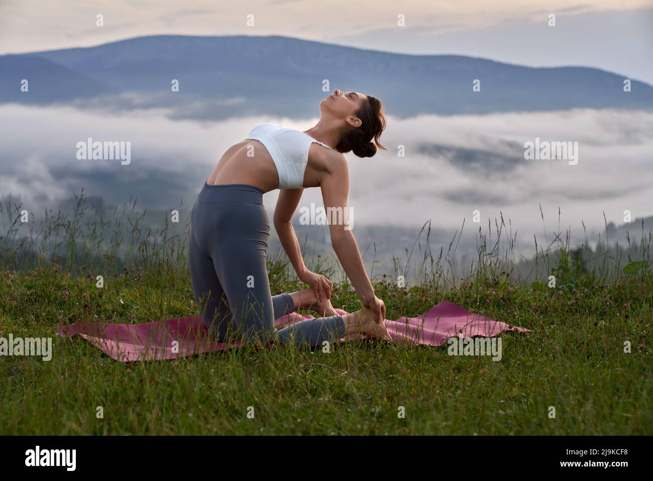 Athletische junge Frau in Sport-BH und Leggings, die den Körper auf einer Yogamatte im Freien dehnt. Weibliche Person, die aktiv in den Sommerbergen trainiert. Stockfoto