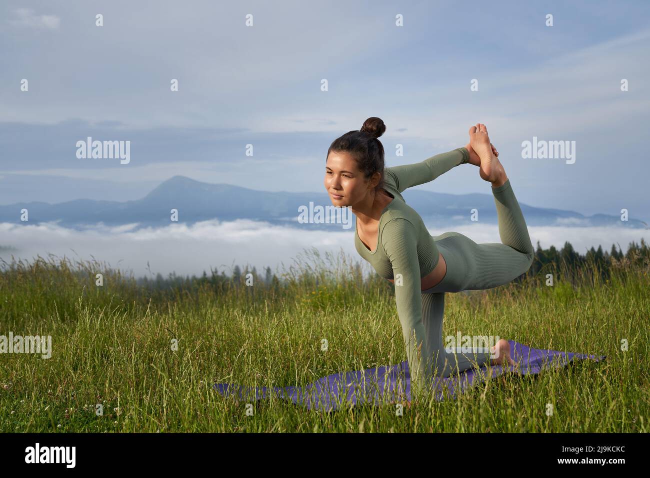 Attraktive Passform Frau in Sportkleidung, die das Gleichgewicht auf einem Knie hält, während sie das Bein im Freien dehnt. Hintergrund von schönen grünen Bergen. Stockfoto