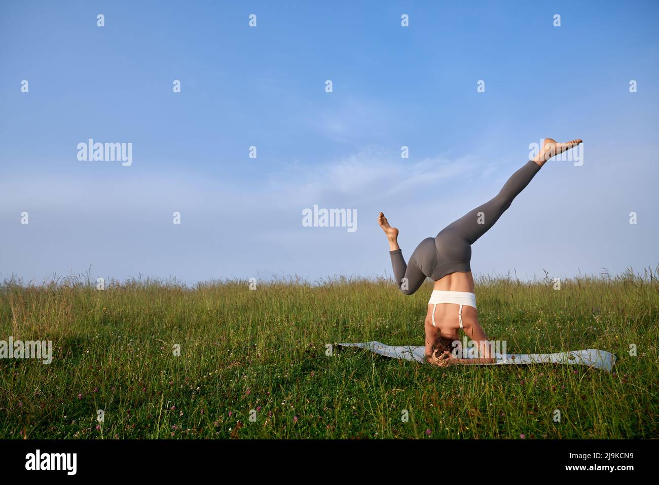 Starke junge Frau im Sportoutfit üben Kopfstand Position zwischen Sommer Berge. Aktive weibliche Person, die während der Morgenstunden im Freien Akro-Yoga macht. Stockfoto