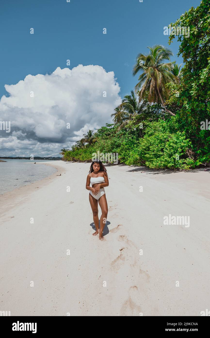 Junge, braune asiatische Frau in einem weißen Bikini an einem tropischen Sandstrand, umgeben von Palmen und Kokospalmen, an einem tropischen sonnigen Tag Stockfoto