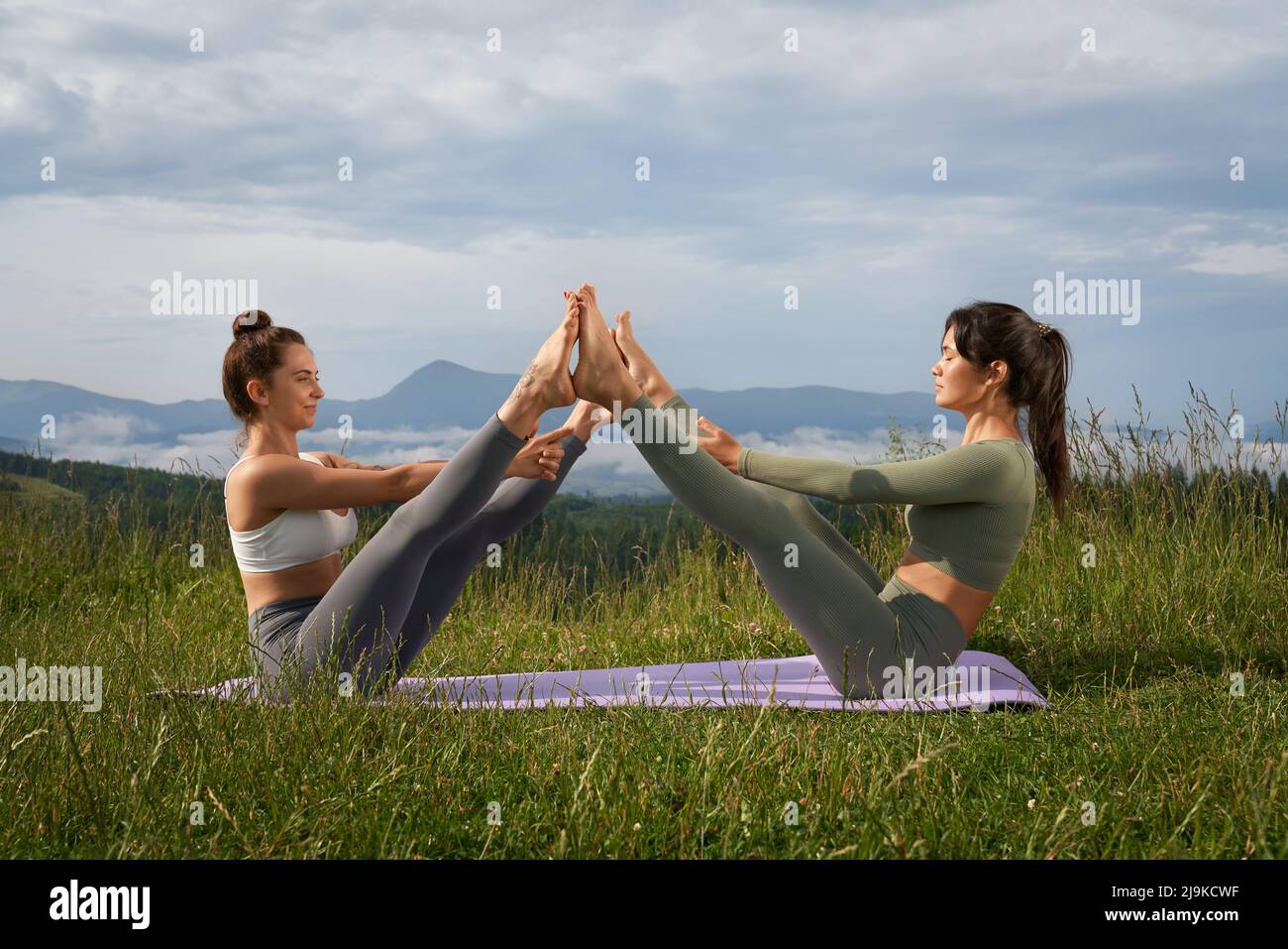 Zwei kaukasische Frauen in aktivem Ohr machen akrobatischen Yoga an frischer Luft. Junge Damen sitzen auf einem Yogamann und strecken die Beine zusammen. Konzept für einen gesunden Lebensstil. Stockfoto