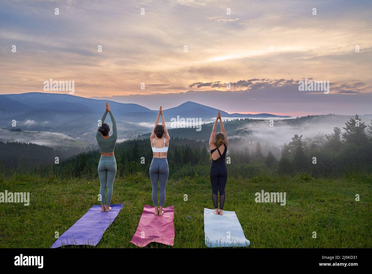 Rückansicht von drei sportlichen Frauen im aktiven Ohr, die im Sommer die Meditation im Freien genießen. Erstaunlicher Sonnenuntergang im Hintergrund. Stockfoto