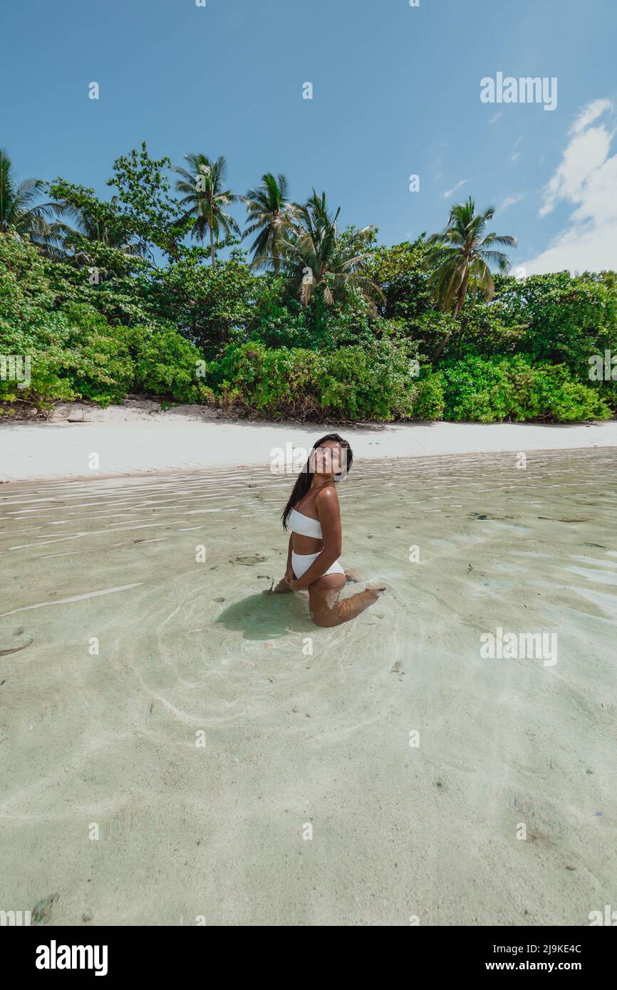 An einem sonnigen Tag in den Tropen kniet ein junges, bräunliches asiatisches Weibchen in einem weißen Bikini auf einem tropischen Sandstrand Stockfoto