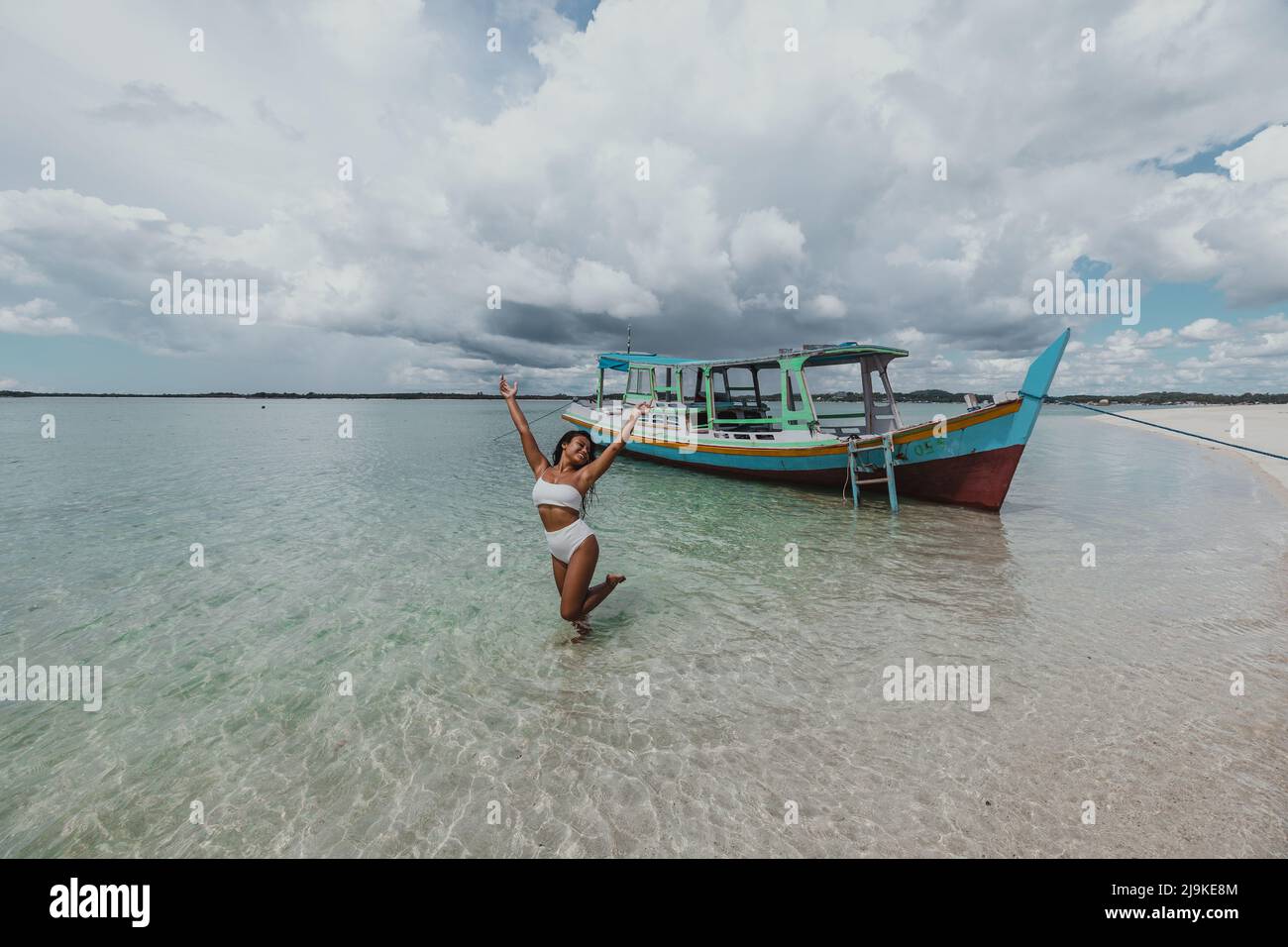 An einem sonnigen Tag mit einem lokalen indonesischen Fischerboot junge, bräunliche asiatische Frau in einem weißen Bikini an einem tropischen Sandstrand Stockfoto