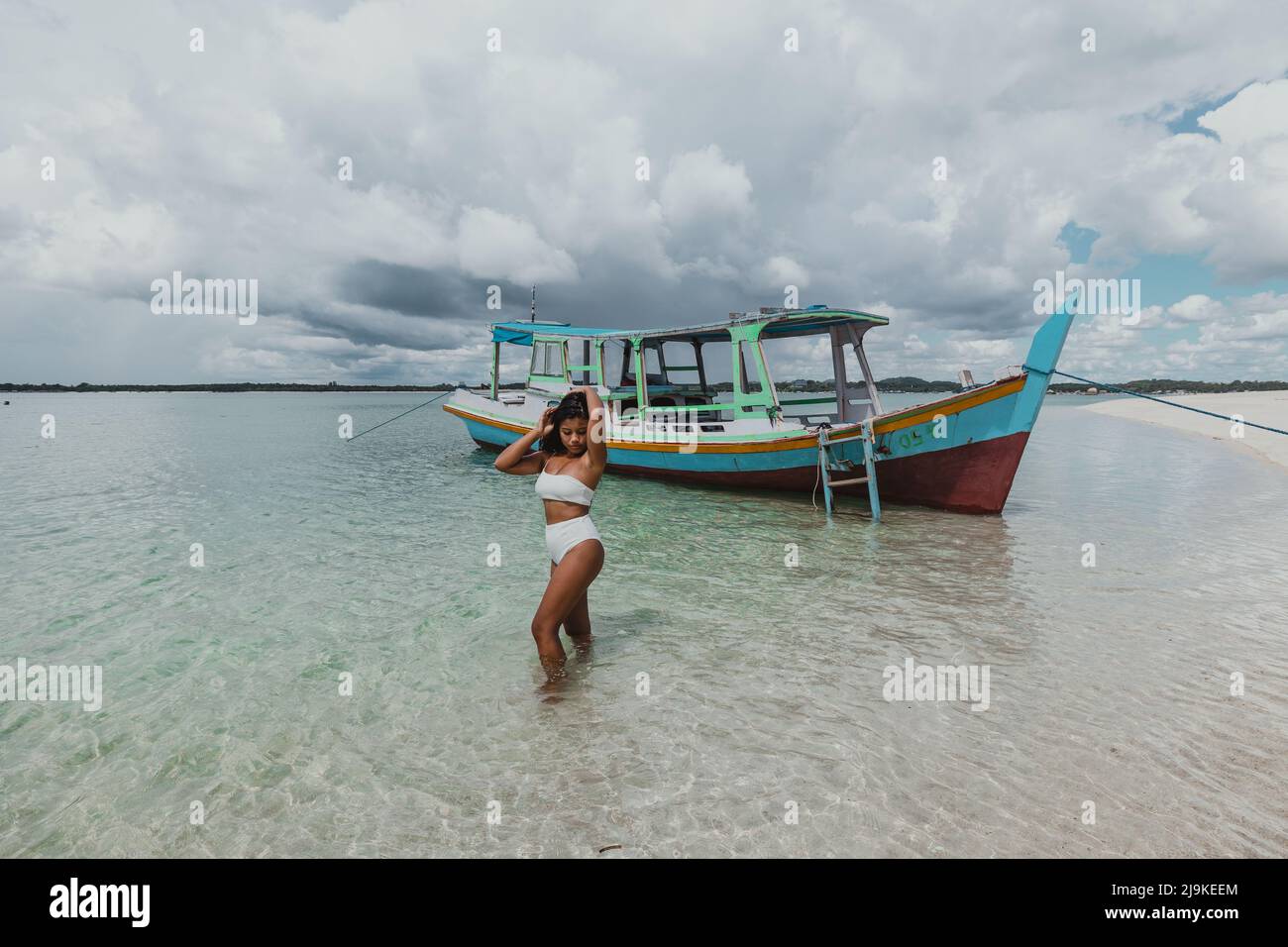 An einem sonnigen Tag mit einem lokalen indonesischen Fischerboot junge, bräunliche asiatische Frau in einem weißen Bikini an einem tropischen Sandstrand Stockfoto