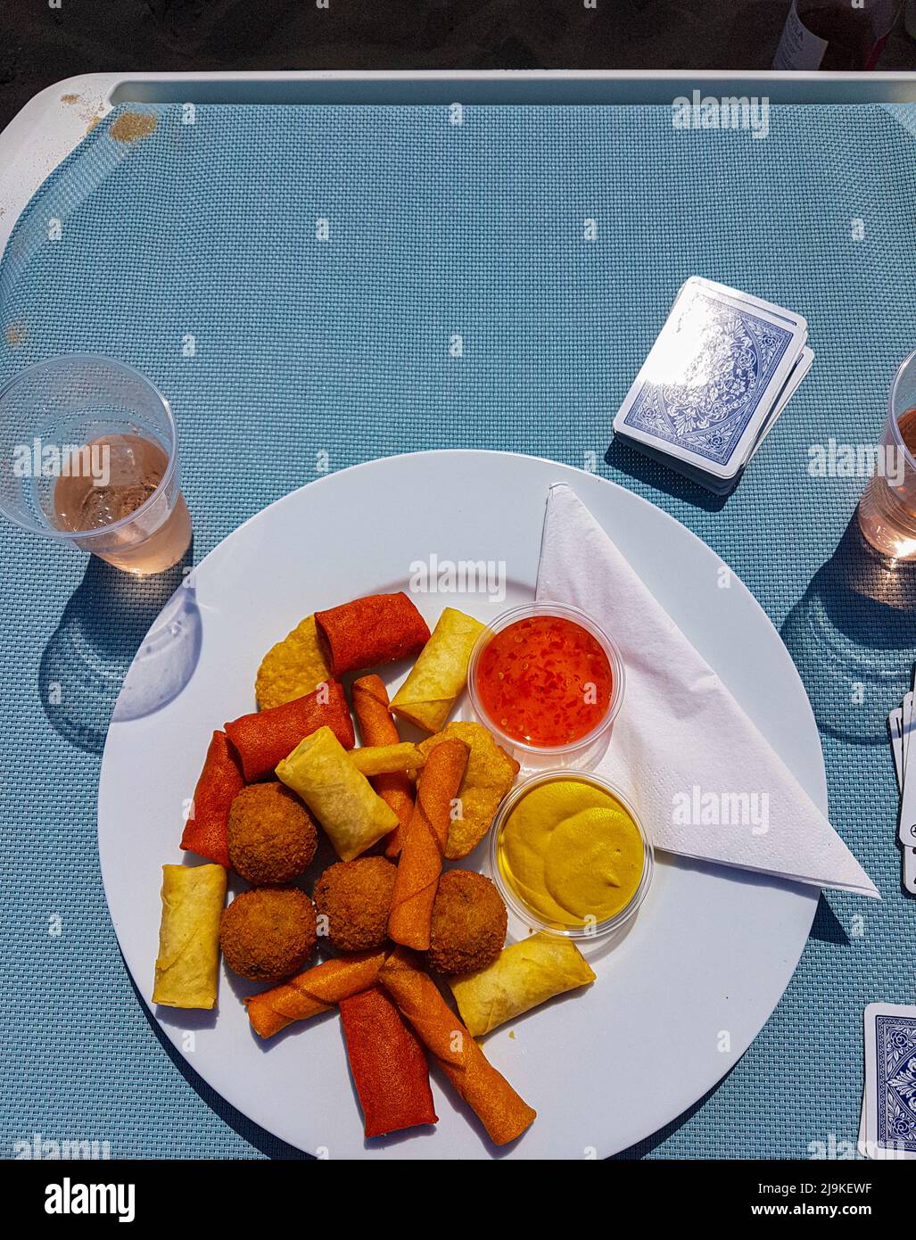 Köstliche verschiedene Snacks auf einem Teller, der sich auf einem Strandbett befindet, mit einer Tasse Rosenwein mit Eis und einem Kartenspiel daneben Stockfoto