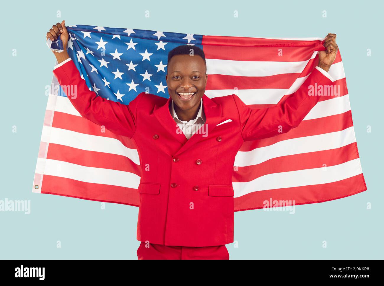 Ein glücklicher, tausendjähriger dunkelhäutiger Mann hebt die amerikanische Nationalflagge auf pastellfarbenem hellblauem Hintergrund. Stockfoto