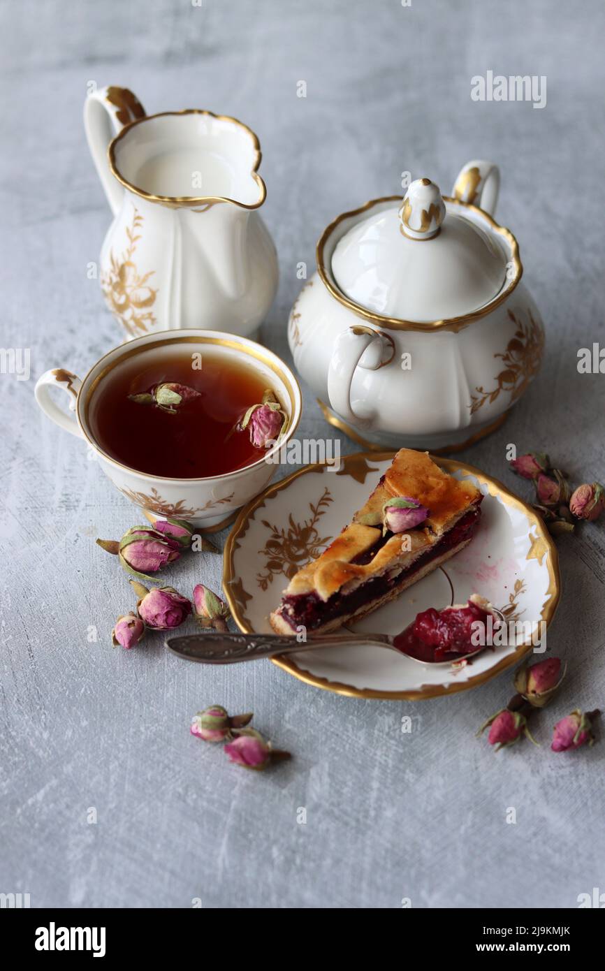 Stillleben mit einer Tasse Tee, einem Krug Milch, einem Kirschkuchen, kleinen Rosenblüten. Weißes Vintage-Geschirr auf einem Tisch. Süßes Frühstück Nahaufnahme Foto. Stockfoto