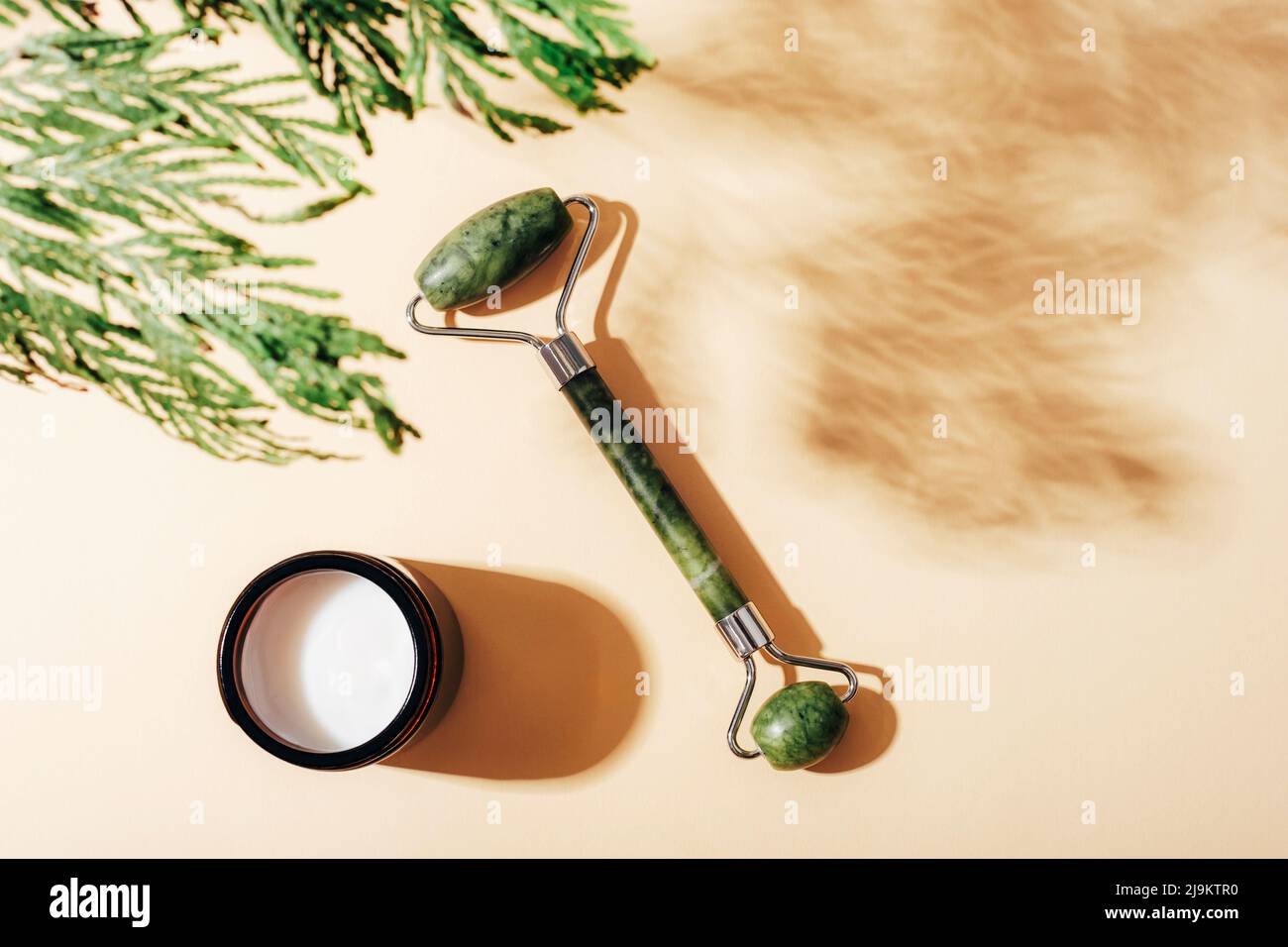 Gesichtswalze und cremefarbenes Glas mit Zypressen-Schatten auf hellbeigem Hintergrund. Naturkosmetik, Aromatherapie, Schönheitsbehandlung Konzept. Draufsicht, Stockfoto
