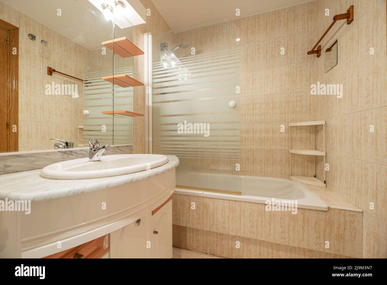 Apartment-WC mit Badewanne mit Glasabtrennung, Schrank mit Spiegel und Lampen, Holzregale und Arbeitsplatte aus weißem und grauem Marmor Stockfoto