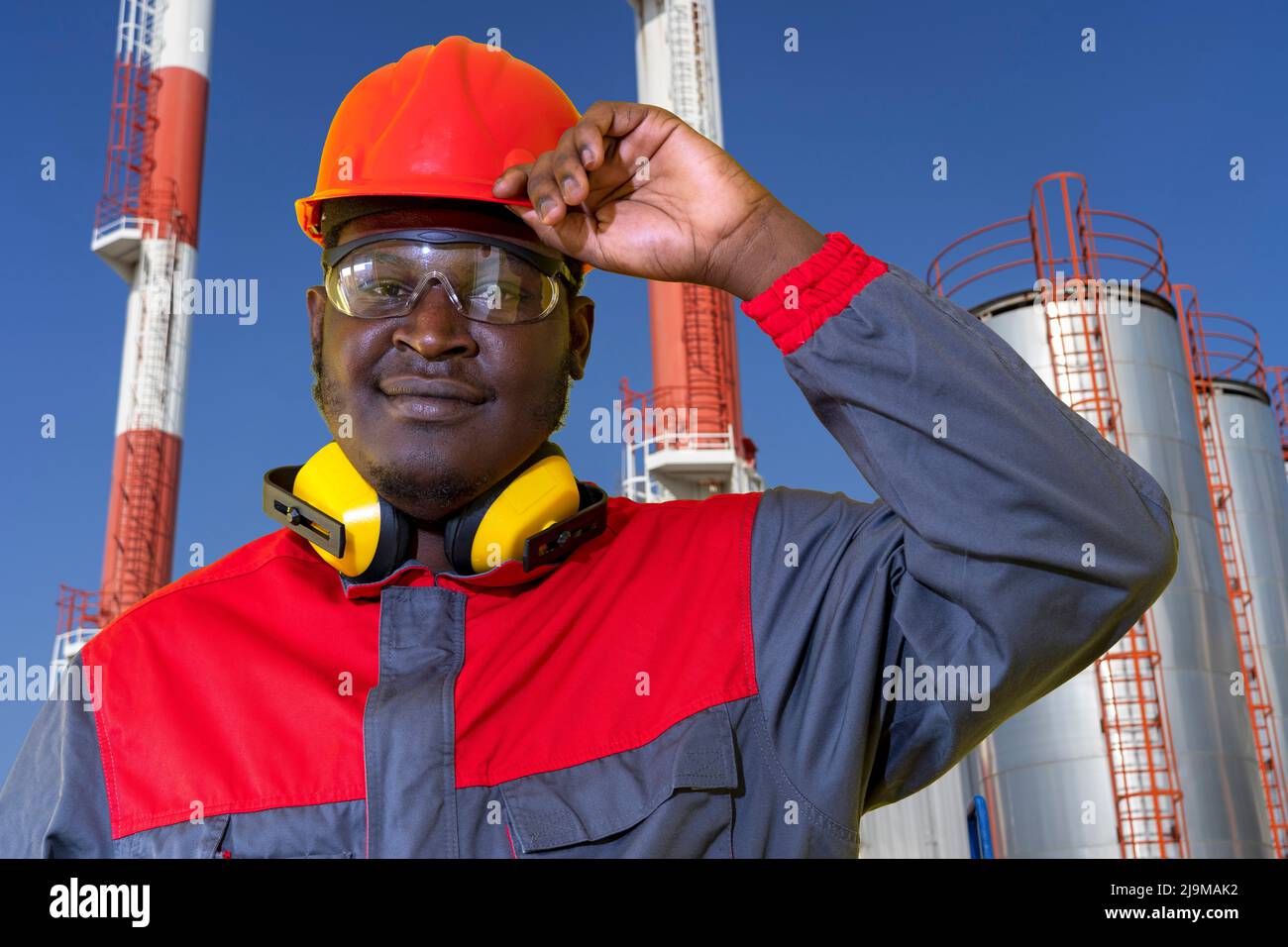 Junge afroamerikanische Ölarbeiterin in Red HardHat steht vor Lagertanks der Ölindustrie. Stockfoto