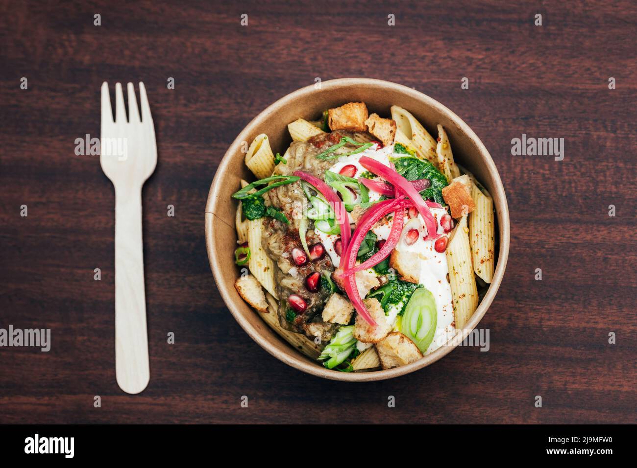 Ansicht von oben auf leckere Pasta in Papierschüssel mit Sauce und Gemüse garniert mit eingelegten Zwiebeln und Granatapfelkernen mit Croutons in der Nähe von Kunststoffgabel serviert Stockfoto