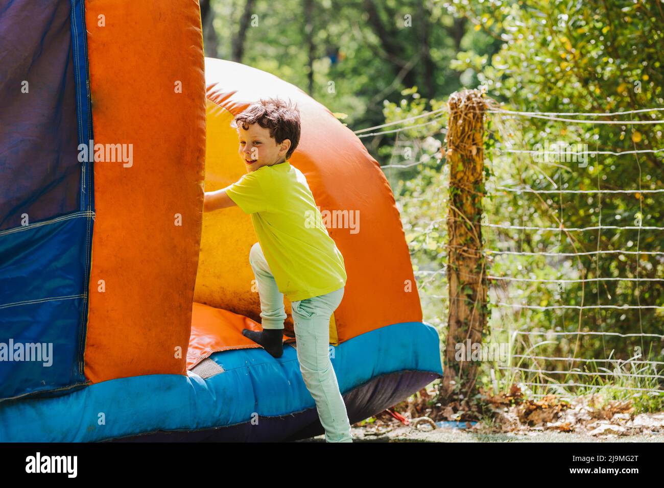 Voller Körper von positiven kleinen Kind mit dunklen Haaren in legerer Kleidung stehen auf weichen aufblasbaren Burg und lächeln auf Spielplatz am sonnigen Sommertag in Stockfoto