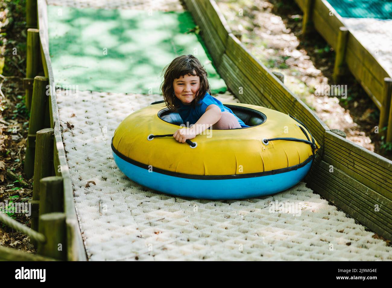 Seitenansicht des entzückten kleinen Kindes im T-Shirt lächelnd, während er auf der Rutsche auf dem Spielplatz im sonnigen Abenteuerpark aufblasbares Schlittenrohr reitet Stockfoto