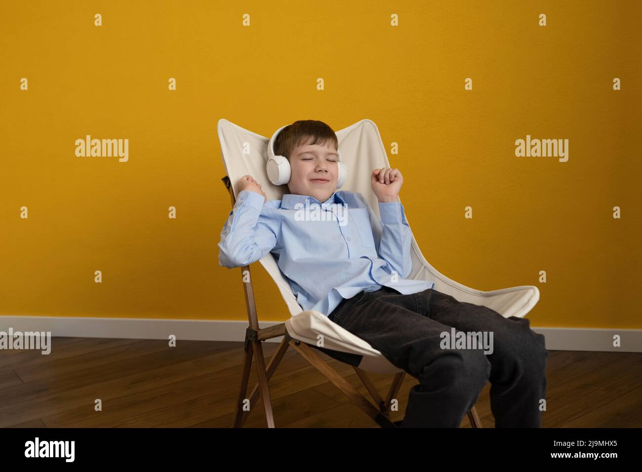 Fröhlicher Junge im blauen Hemd, der die Augen schließt und tanzt, während er auf dem Liegestuhl an der gelben Wand sitzt und Musik in kabellosen Kopfhörern hört Stockfoto