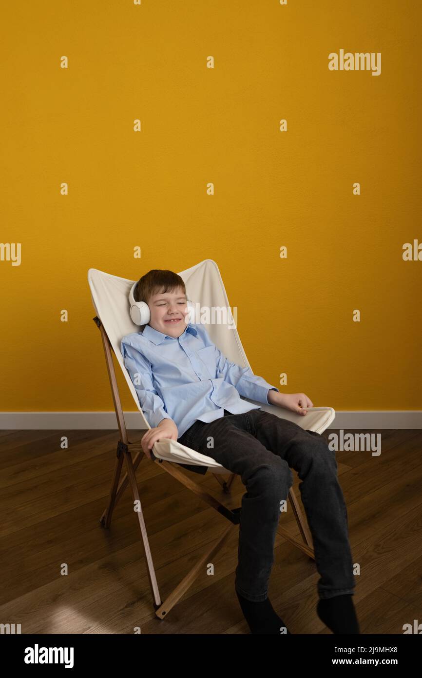 Fröhlicher Junge im blauen Hemd, der die Augen schließt und tanzt, während er auf dem Liegestuhl an der gelben Wand sitzt und Musik in kabellosen Kopfhörern hört Stockfoto