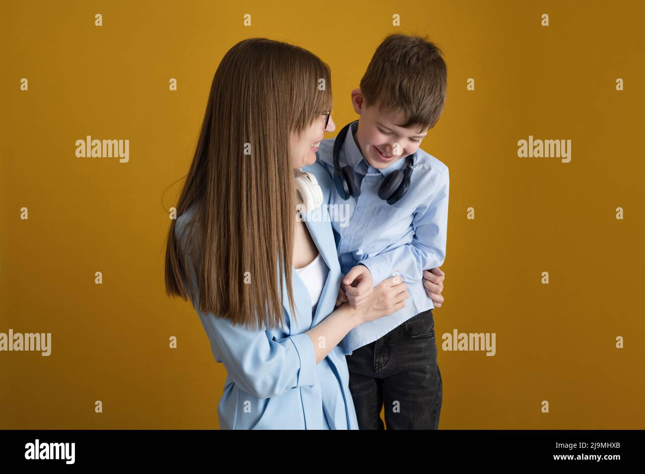 Glückliche Erwachsene Mutter mit langen Haaren umarmt und kitzelt kichernden Sohn, während Spaß zusammen vor gelbem Hintergrund Stockfoto