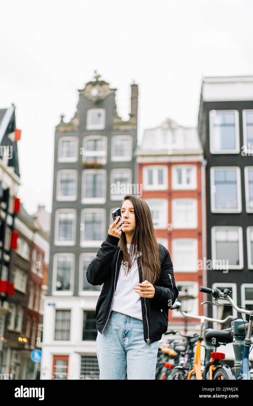 Niedriger Winkel der ernsthaften jungen ethnischen weiblichen Touristen mit langen dunklen Haaren in trendigen Outfit Aufnahme Sprachnachricht über Handy, während sich dagegen Stockfoto