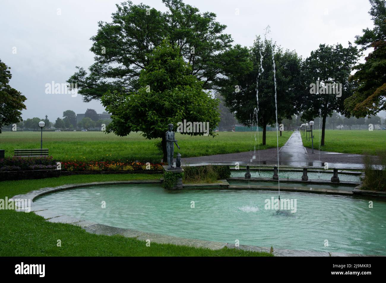 Blick auf einen wunderschönen Teich mit einer Statue und einem Brunnen im Hohematte Park an der Hauptstraße von Interlaken, erfasst an einem regnerischen Tag. Stockfoto