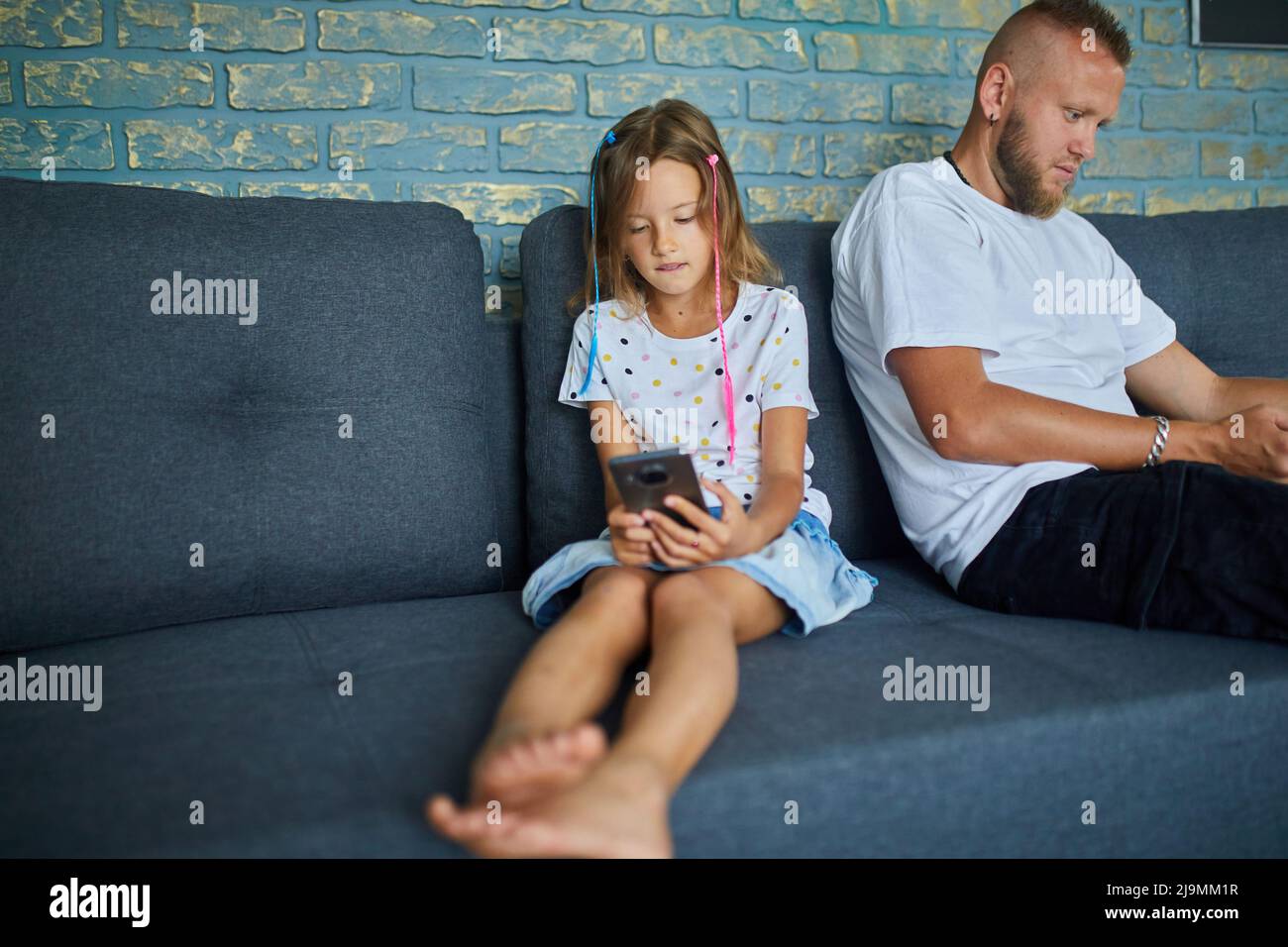 Vater und Tochter stöbern im Mobiltelefon, während sie sich zu Hause auf dem Sofa entspannen, Frau und Mädchen spielen auf Smartphones, während sie sich wochenlang auf der Couch ausruhen Stockfoto