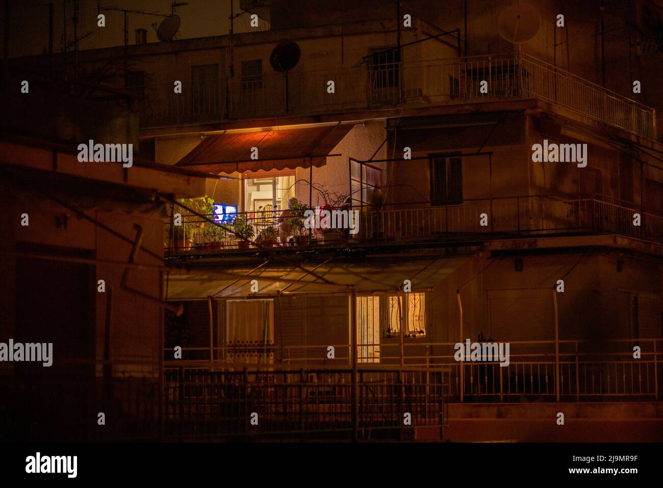 Mann auf dem Balkon, der nach Einbruch der Dunkelheit in Athen allein Fernsehen schaute Stockfoto