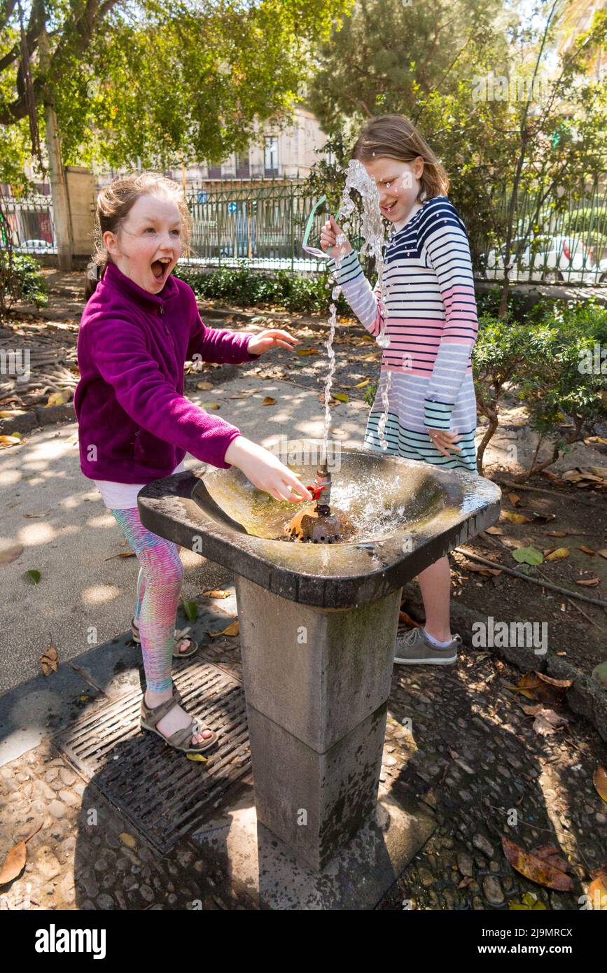 Junge Mädchen überraschten den hohen Druck eines Wasserbrunnens, als sie sich darauf vorbereitete, ein Getränk Wasser / Trinken aus einem durstlöschenden öffentlichen Brunnen draußen, in einem Stadtpark, in Catania, Sizilien, zu nehmen. Italien. (129) Stockfoto