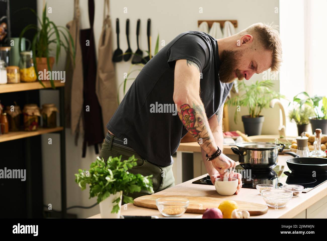 Hübscher junger Kaukasischer mit Tätowierungen auf Armen, der in der Küche steht und Gewürze mit Mörser und Stößel zermalmt Stockfoto