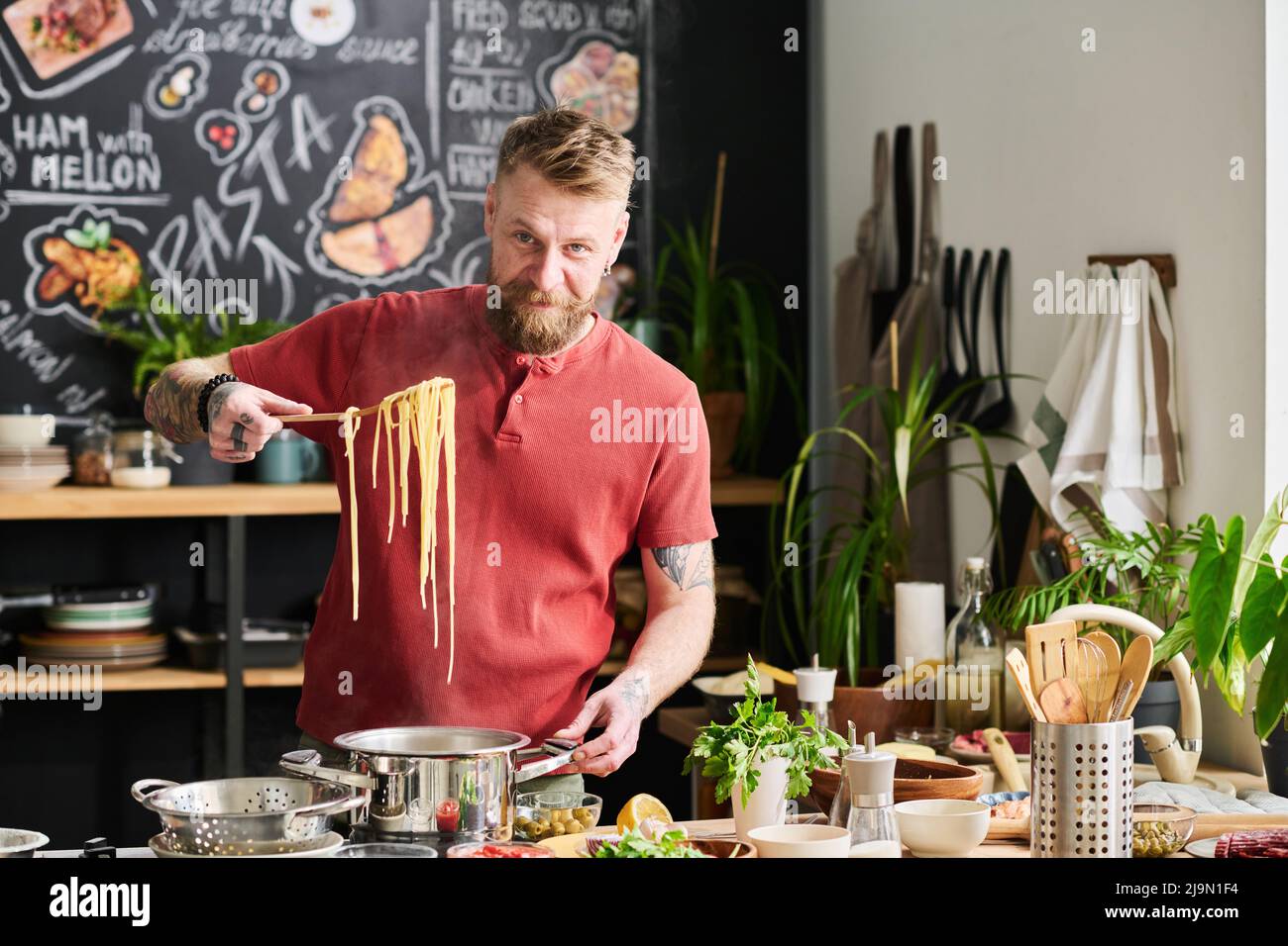 Fröhlicher kaukasischer Mann mit Bart im Gesicht, der Nudeln aus heißem Wasser für Pasta nimmt und dabei die Kamera anschaut Stockfoto