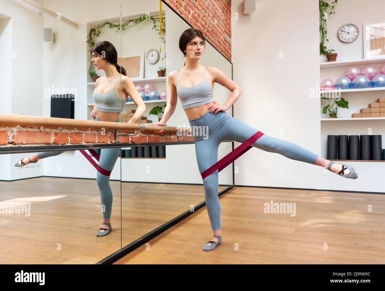 Ganzer Körper der schlanken Tänzerin, die in der Nähe von Barre steht und mit Gummiband trainiert, während sie Kniebeugen macht und im Tanzstudio mit Spiegel wegschaut. Stockfoto
