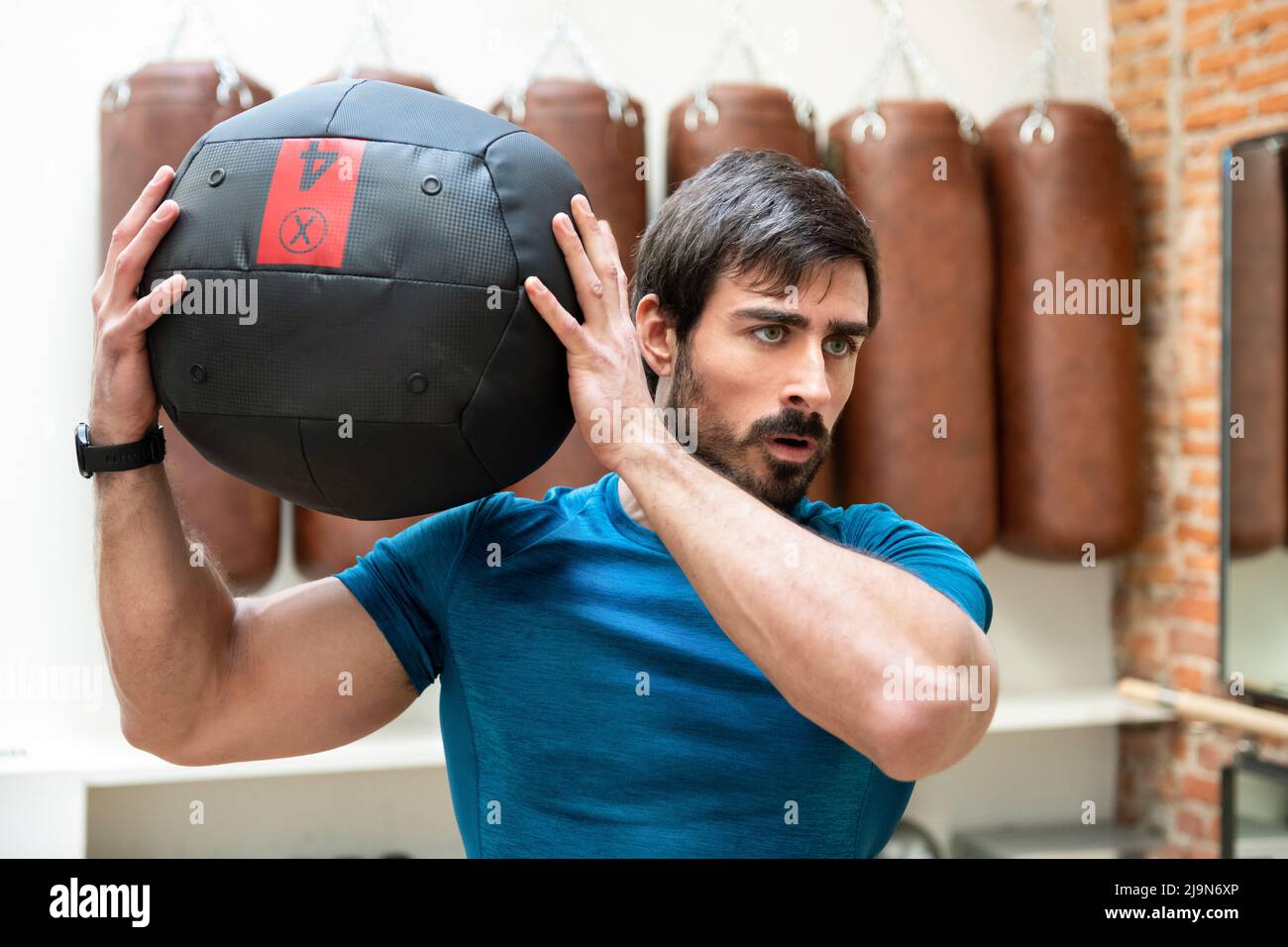Entschlossener, athletischer Mann mit muskulösem Körper, der während des intensiven Trainings im Fitnessstudio mit schwerem Medizinball trainiert Stockfoto