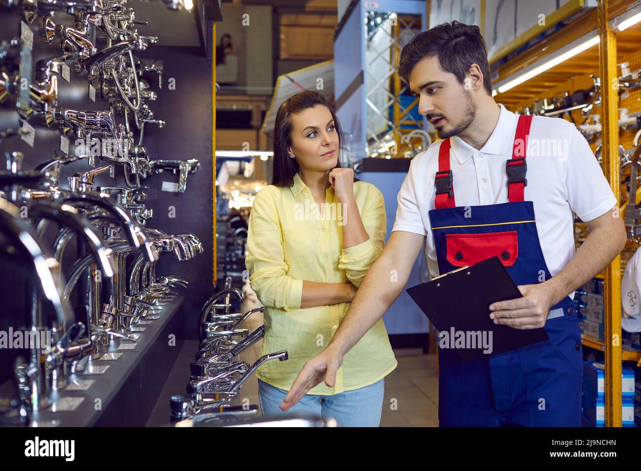 Junge Frau mit langen Haaren, die einen Wasserhahn für Bad oder Küche in einem Einrichtungsgeschäft mit einem Berater wählt. Stockfoto