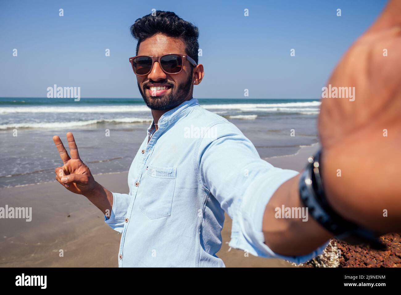 Junger afroamerikanischer Mann in blauem Hemd und Jeans, der ein Selfie-Portrait auf der Frontkamera eines Smartphones am Meeresstrand macht. Stockfoto