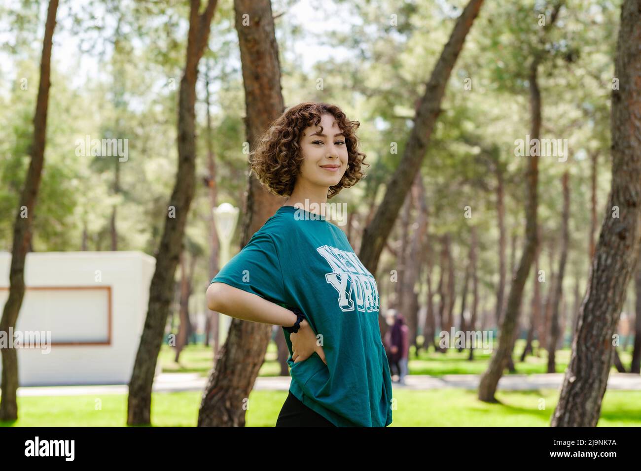 Seitenansicht einer jungen Rotschopf-Frau in einem grünen T-Shirt, die auf dem Stadtpark steht, im Freien die Hände an den Hüften hält und mit großem Lächeln auf die Kamera blickt. Sm Stockfoto