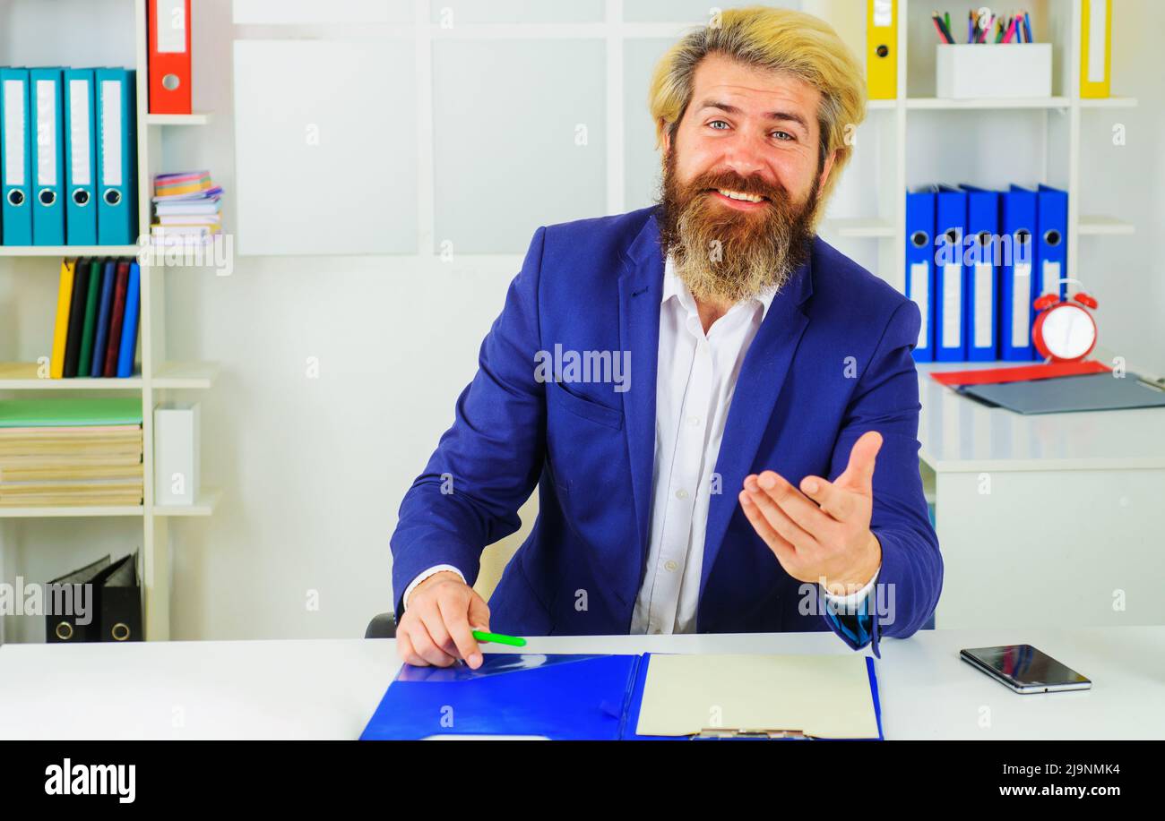 Im modernen Büro sitzt ein stilvoller, lächelnder Geschäftsmann im Anzug am Schreibtisch. Unternehmenswachstum. Führung. CEO. Stockfoto