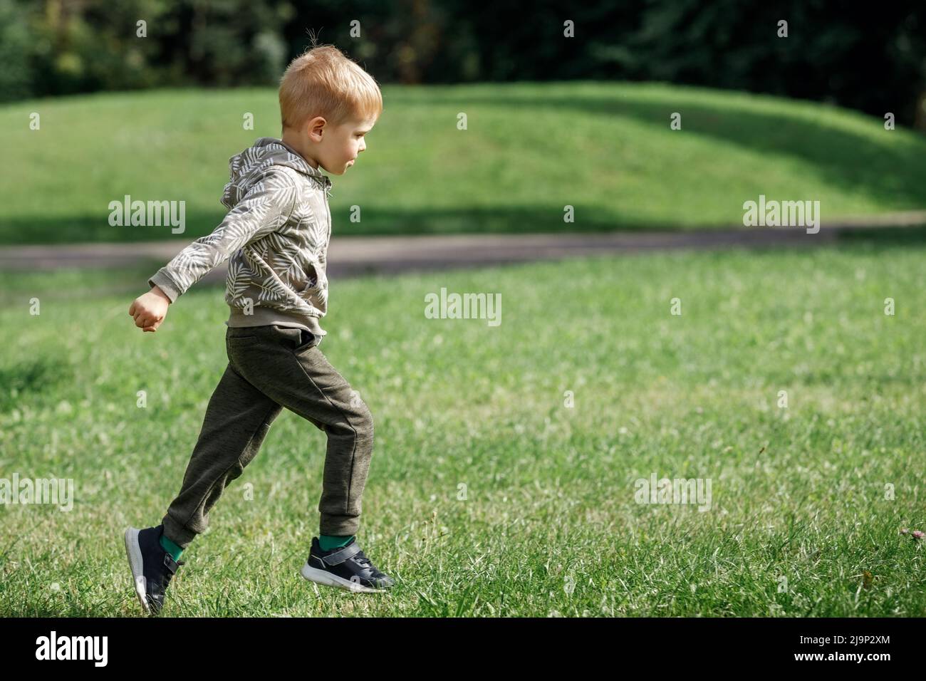 Netter Junge, der über Gras läuft und lächelt. Ein agiles, glückliches Kind spielt in einem Sommer-Stadtpark Sport. Stockfoto