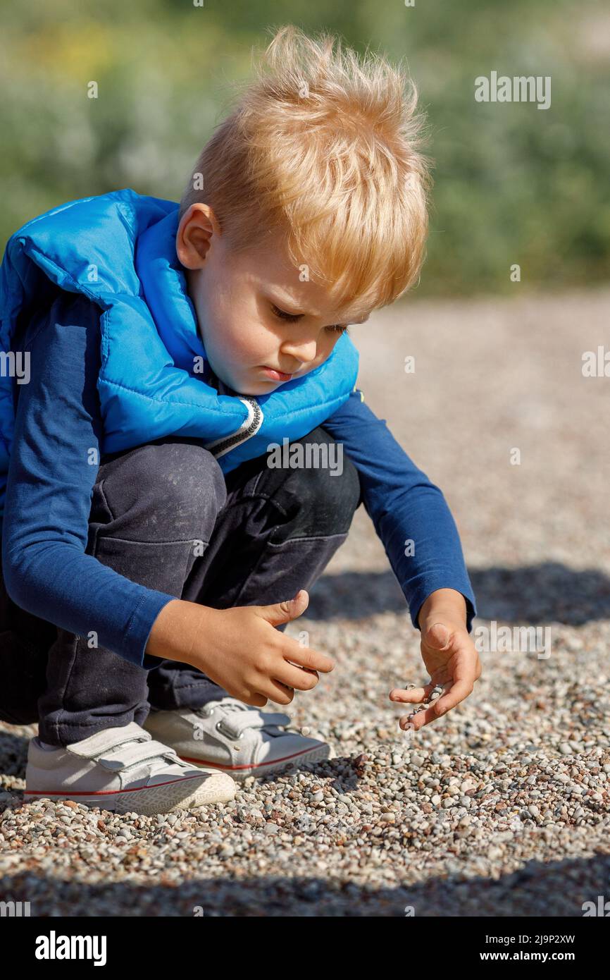 Ein kleiner Junge mit einer blauen Weste spielt draußen mit kleinen Kieselsteinen. Das Kind erkundet den Boden mit seinen eigenen Händen, er ist sehr an kleinen Kebb interessiert Stockfoto
