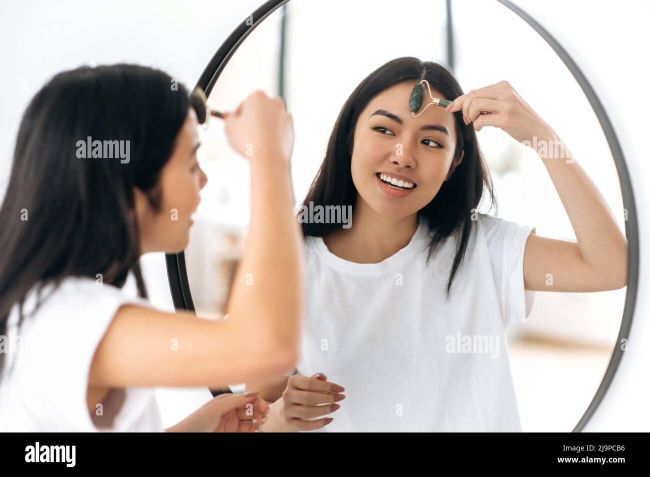 Happy chinese Brunette hübsches Mädchen in weißem T-Shirt, steht vor einem Spiegel zu Hause, macht vorsichtig ihre morgendliche Schönheitsroutine, kümmert sich um ihre Gesichtshaut, verwendet eine Gesichtssteinrolle, lächelnd Stockfoto