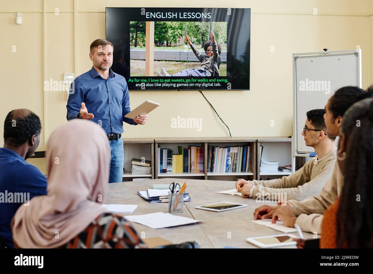 Professioneller Englischlehrer, der vor multiethnischen Migrantenstudenten am Tisch steht und ihnen etwas erklärt Stockfoto