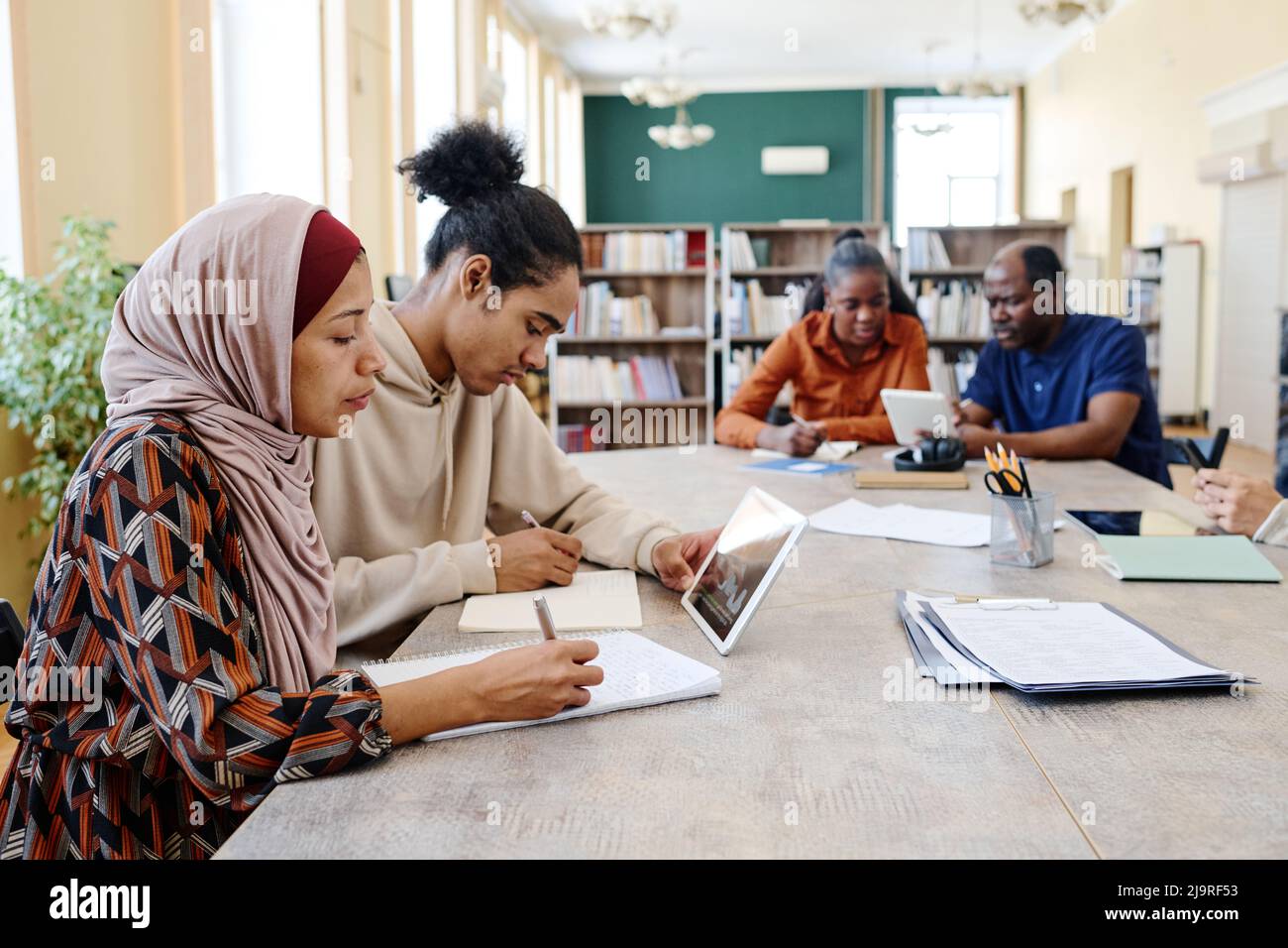 Eine Gruppe ethnisch diverser Studenten, die am Tisch sitzen und Bildungsvideos auf einem digitalen Tablet ansehen und Schreibaufgaben erledigen Stockfoto