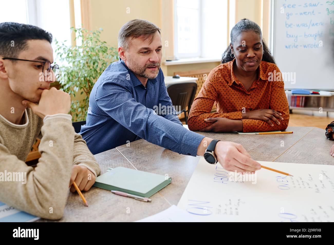 Reifer Lehrer, der mit eingewanderten Schülern arbeitet und Bleistift auf das auf dem Poster geschriebene Wort zeigt und es erklärt Stockfoto