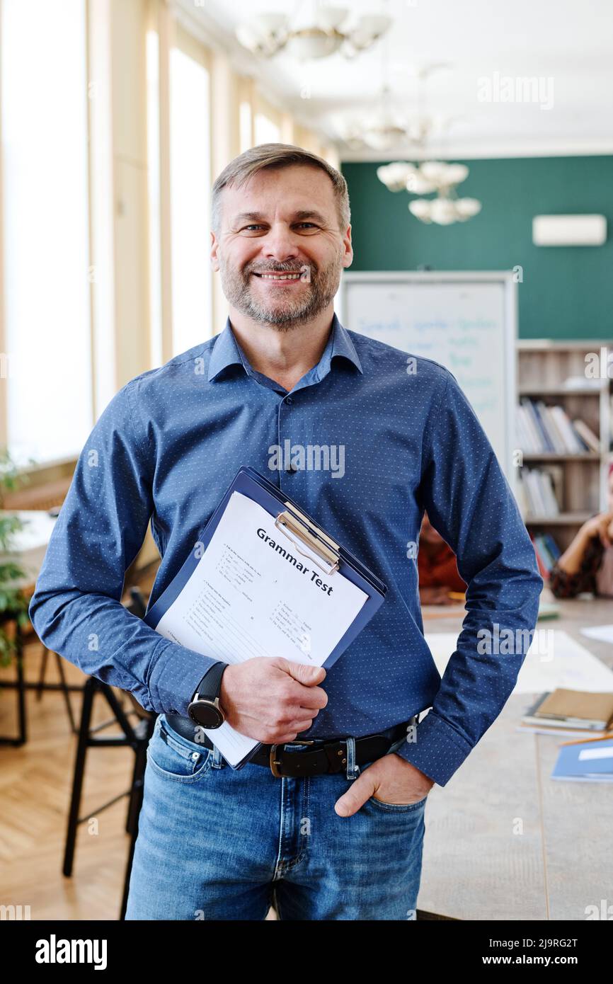 Vertikale mittlere Aufnahme von freudigen reifen kaukasischen Mann Unterricht Englisch Sprache halten Zwischenablage mit Grammatiktests lächeln an der Kamera Stockfoto