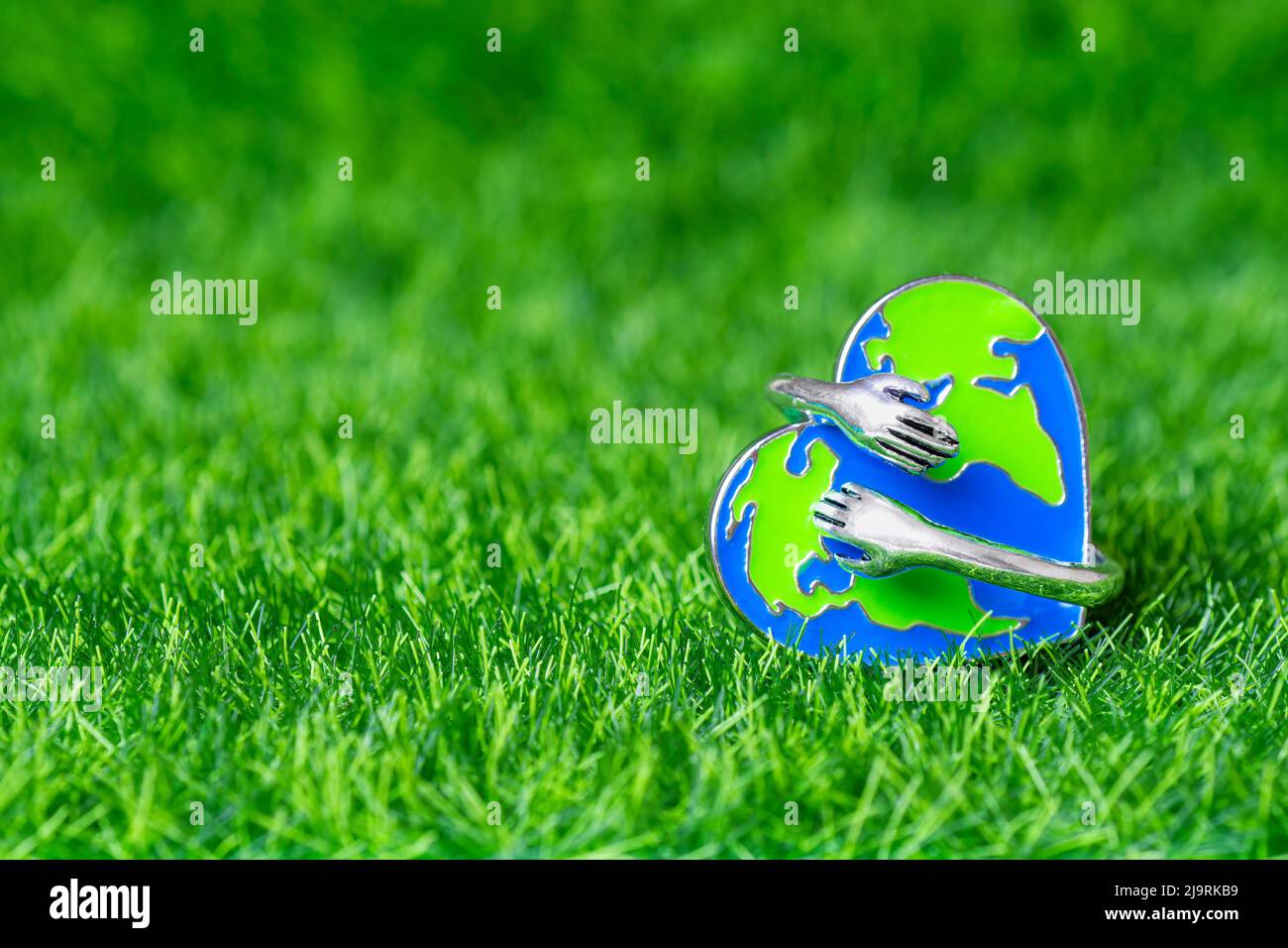 Nahaufnahme einer emaillierten Nadel in Form von kleinen Händen, die einen herzförmigen Globus umarmen, der auf einem grünen Grashintergrund mit Kopierfläche platziert ist. Umweltschutz Stockfoto