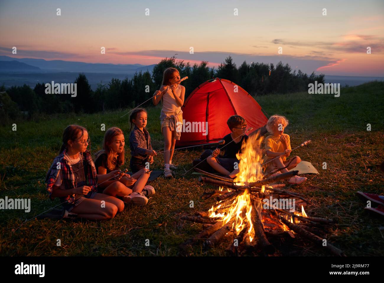 Seitenansicht einer Gruppe von Kindern, die auf dem Campingplatz in den Bergen essen. Sechs Kinder beim Abendessen, Camping in der Nähe des Kamins mit rotem Zelt auf dem Hintergrund. Konzept des Reisens. Stockfoto