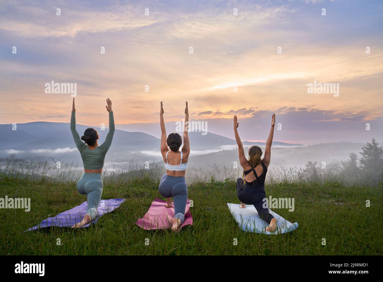 Rückansicht von Baumfrauen, die auf Gras als Krieger posieren, Yoga in den Bergen praktizieren. Virabhadrasana mit den Händen, die zum Himmel rasen Konzept des gesunden Rückzugs. Stockfoto